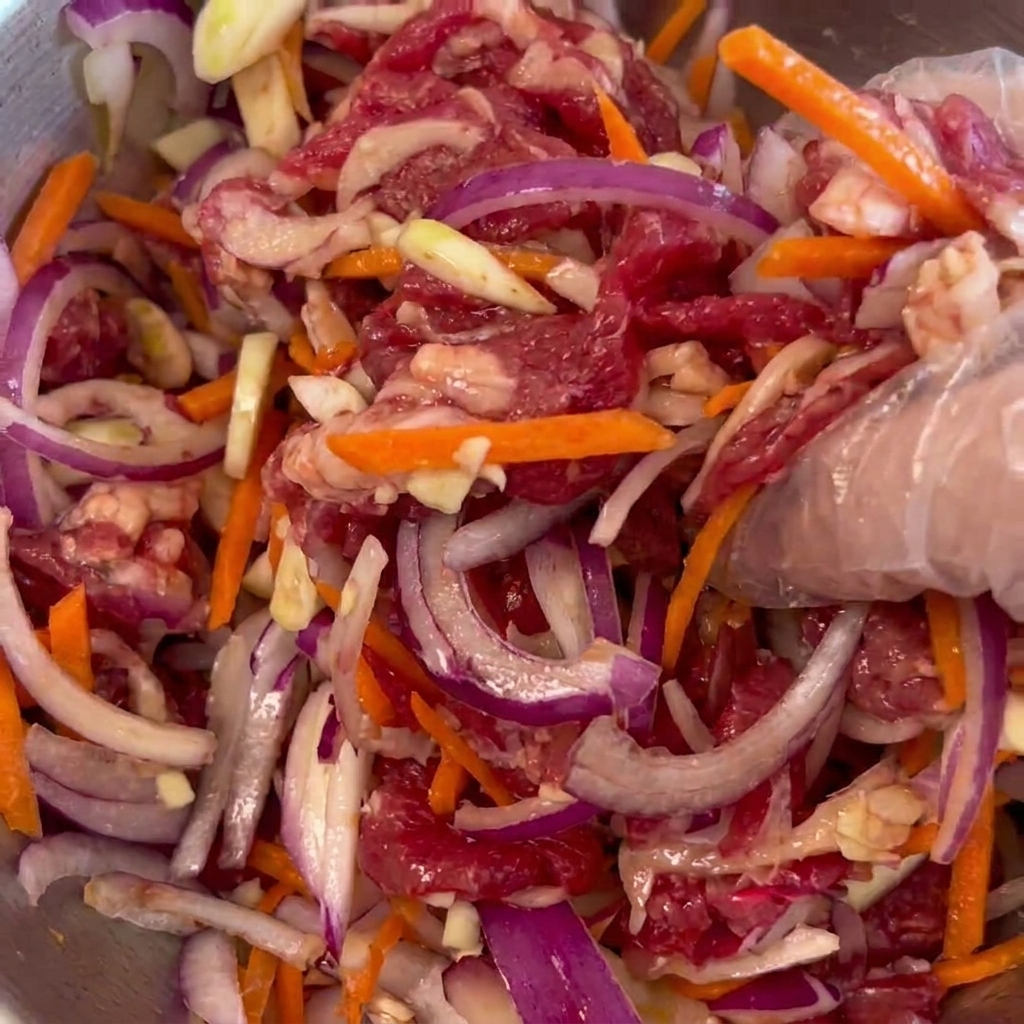 Una mano con guante mezclando la carne de ternera, la cebolla morada y la zanahoria en un bol metálico.