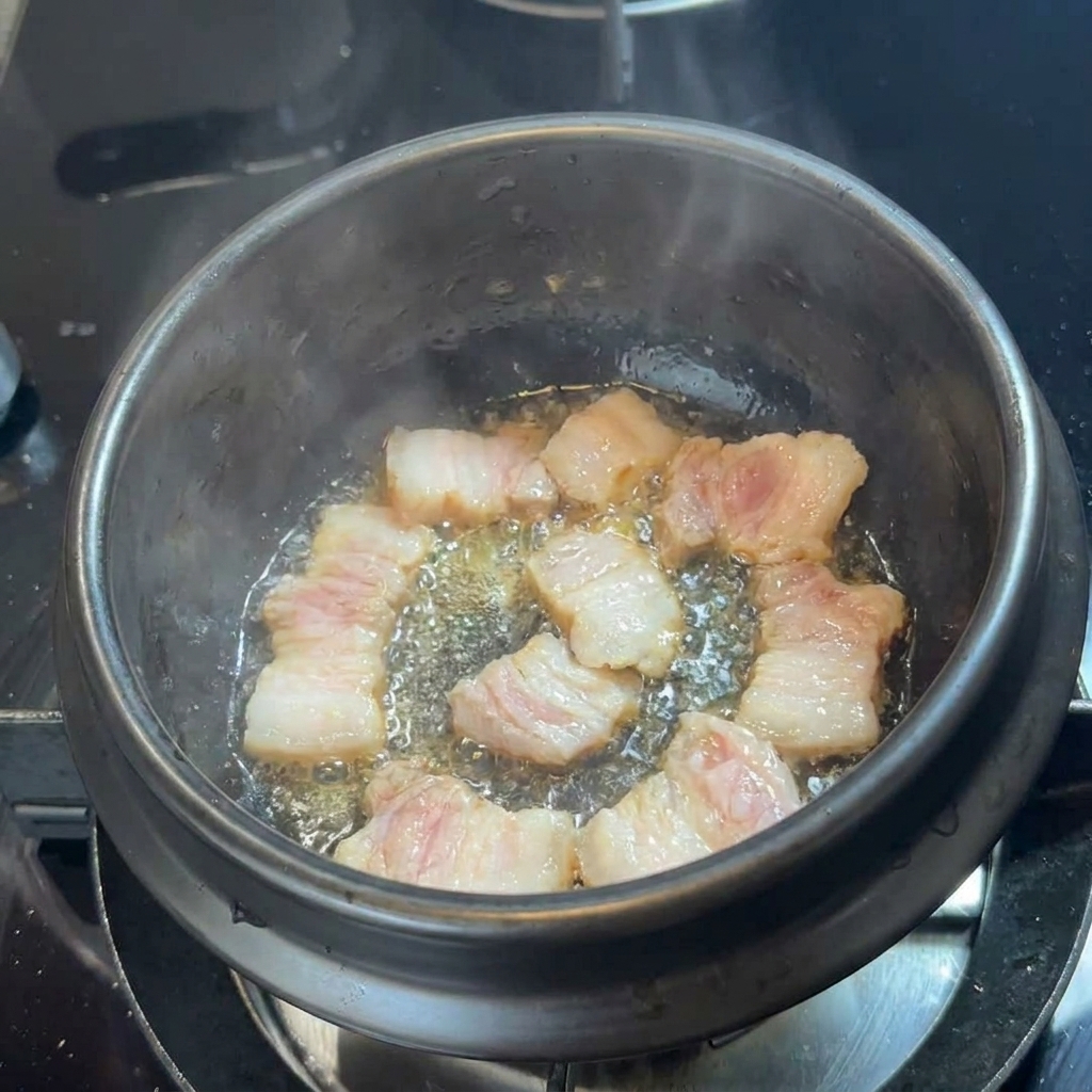 Tiras de panceta de cerdo dorándose en su propia grasa en el fondo de una cazuela negra.
