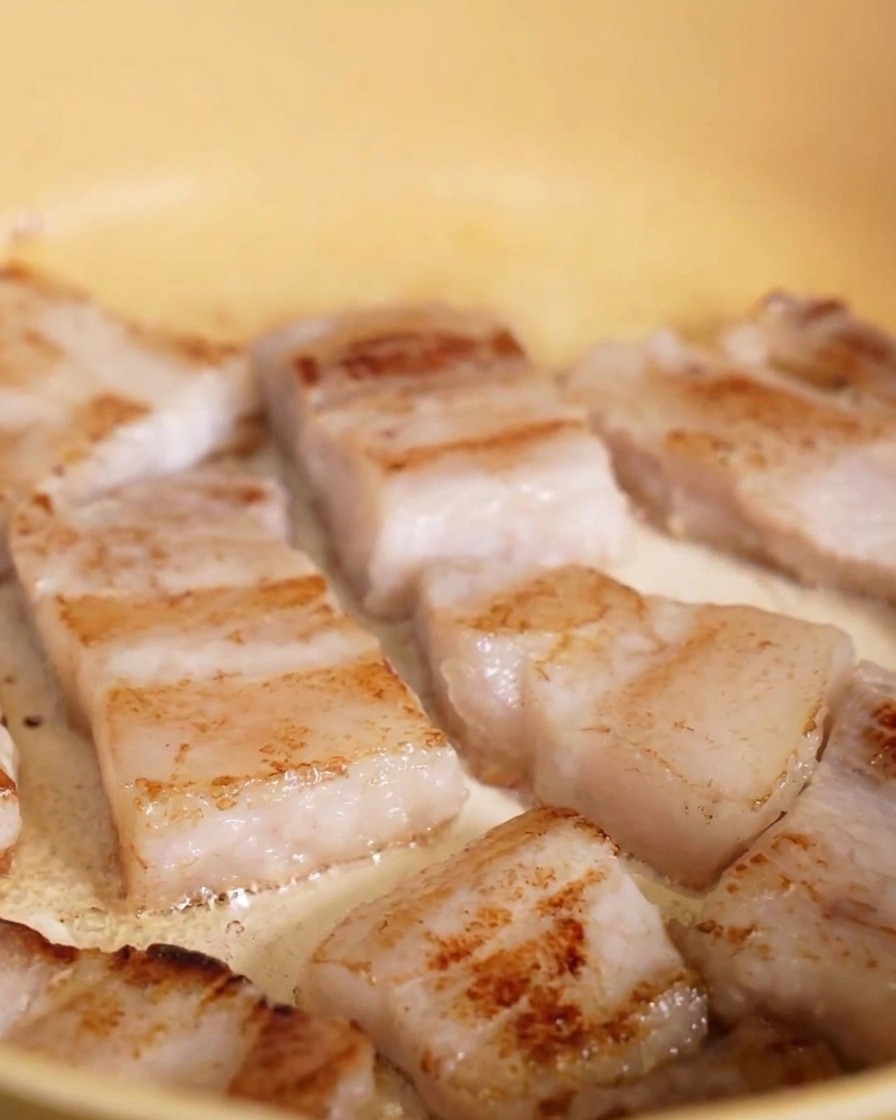 Lonchas de panceta de cerdo friéndose en una sartén amarilla, con la parte inferior mostrando una costra dorada.