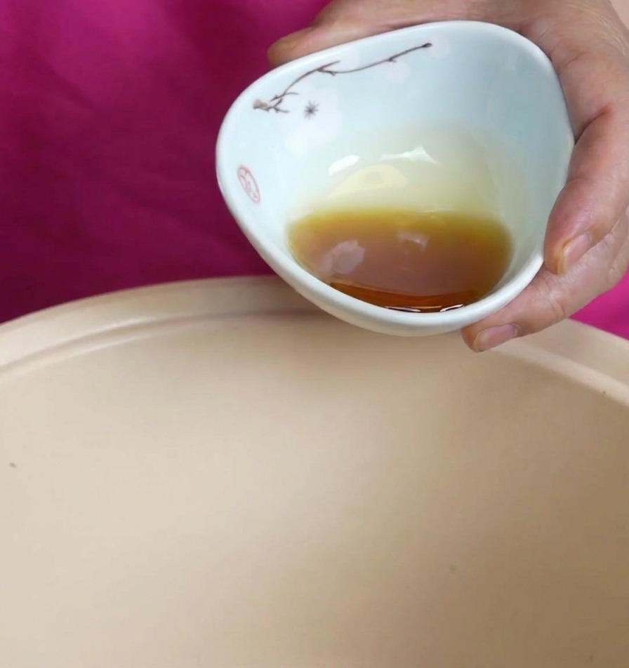 Aceite de sésamo de color ámbar rico vertiéndose desde un pequeño bol de cerámica en una olla de cocción beige.