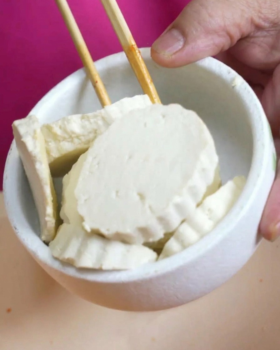 Un pequeño bol blanco lleno de rodajas gruesas de tofu blanco fresco sostenido sobre una olla de sopa.