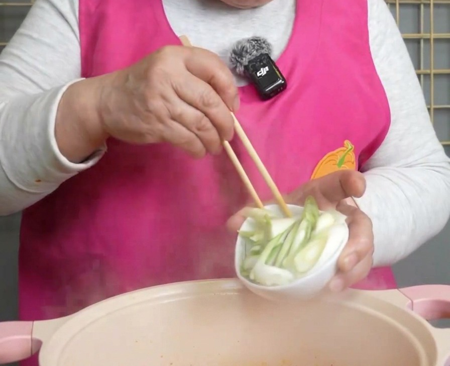 Cebolletas frescas recién picadas vertiéndose desde un pequeño bol blanco en una olla humeante de sopa de kimchi roja.