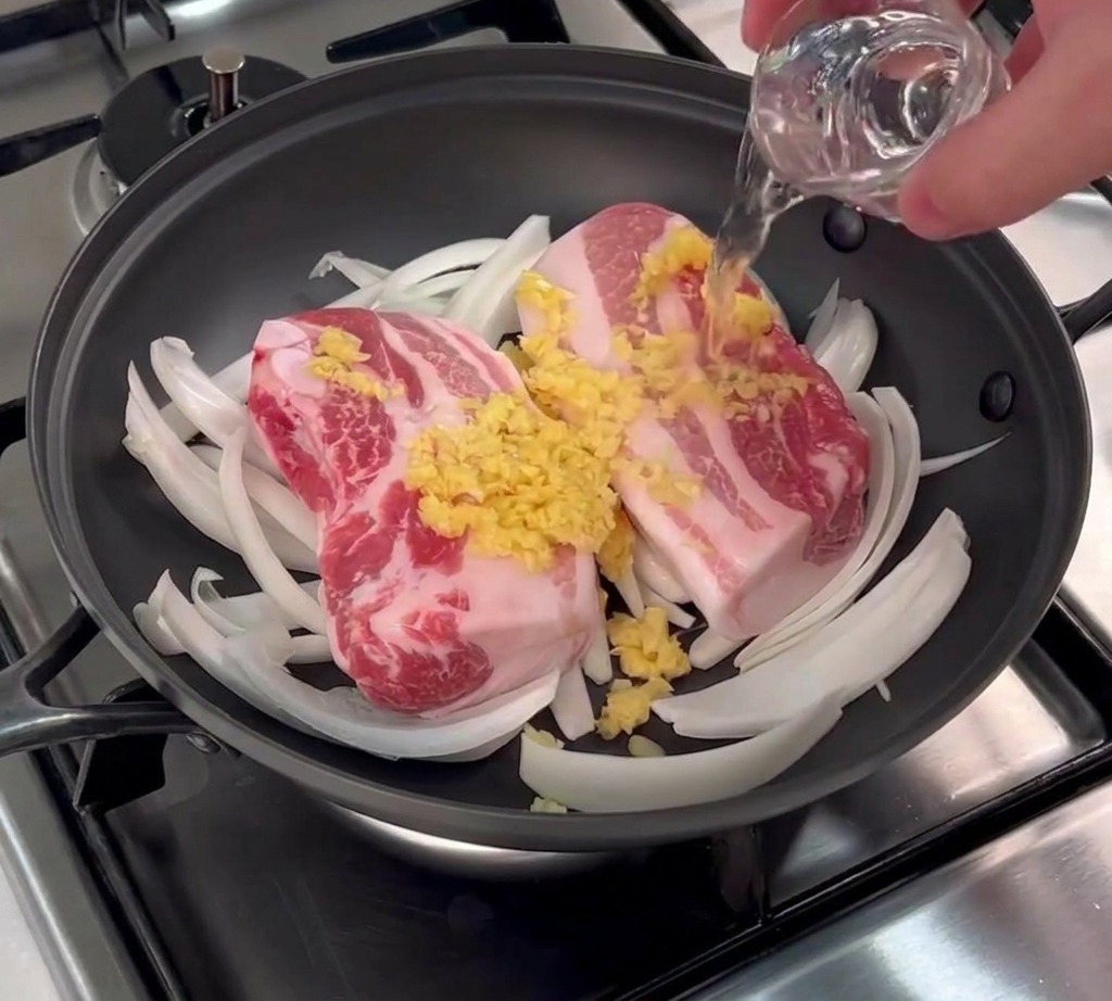 Vino de arroz transparente vertiéndose sobre panceta cruda con ajo picado en una sartén negra.