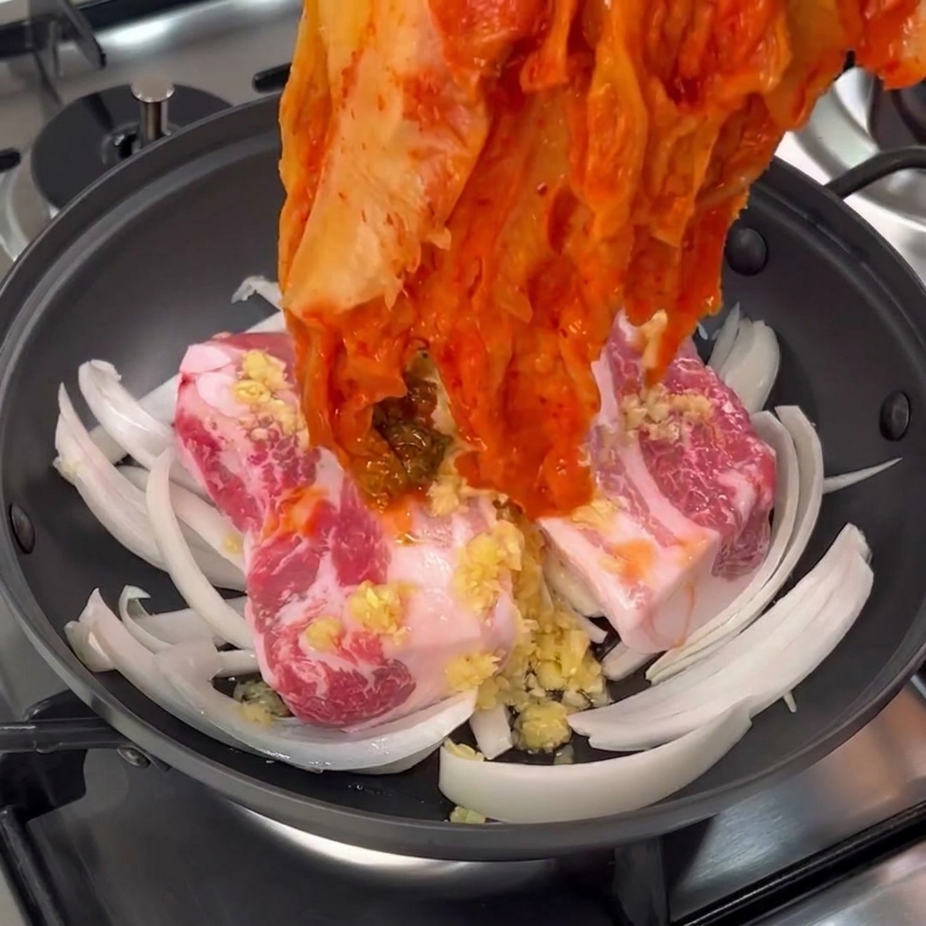 Una mano colocando un fajo de kimchi de col roja fermentada sobre trozos de panceta cruda.