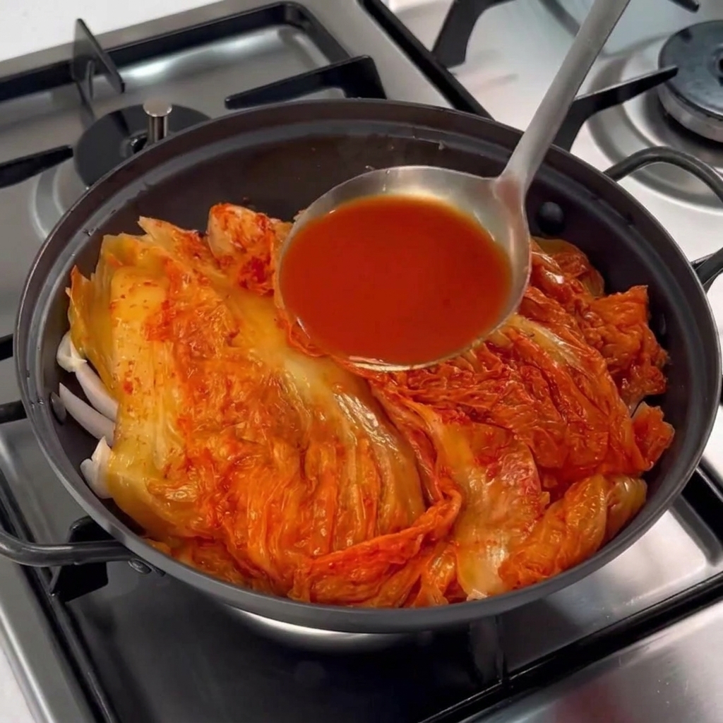 Un cucharón de metal vertiendo salmuera de kimchi de color rojo vibrante sobre la olla.