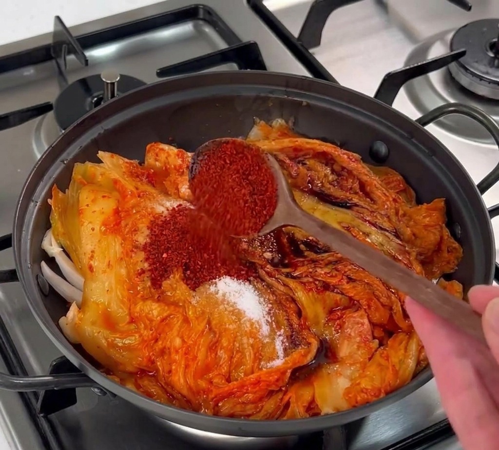 Cuchara de madera vertiendo copos de chile rojo coreano sobre una capa de kimchi fermentado.