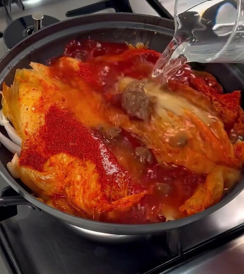 Agua clara vertiéndose desde una jarra de cristal en una olla con kimchi, panceta y especias.