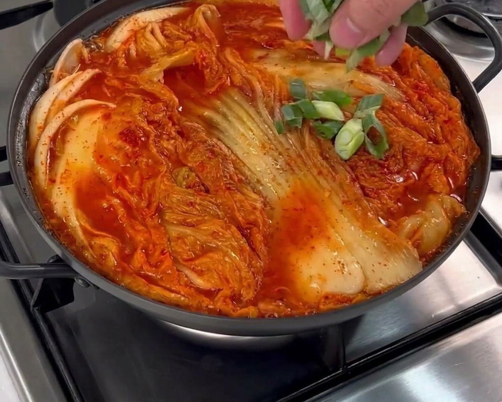 Una mano esparciendo cebolletas frescas picadas sobre la olla llena de caldo rojo.