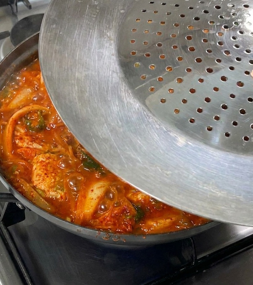 Mano colocando una tapa metálica sobre una sartén burbujeante de estofado de kimchi en la estufa.