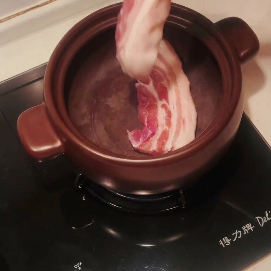 Tiras de panceta de cerdo cruda colocadas en una olla de barro oscura precalentada con pinzas metálicas.