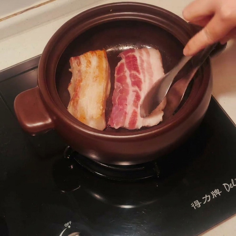 Tiras de panceta en una olla de barro siendo volteadas con pinzas, mostrando un lado perfectamente dorado.