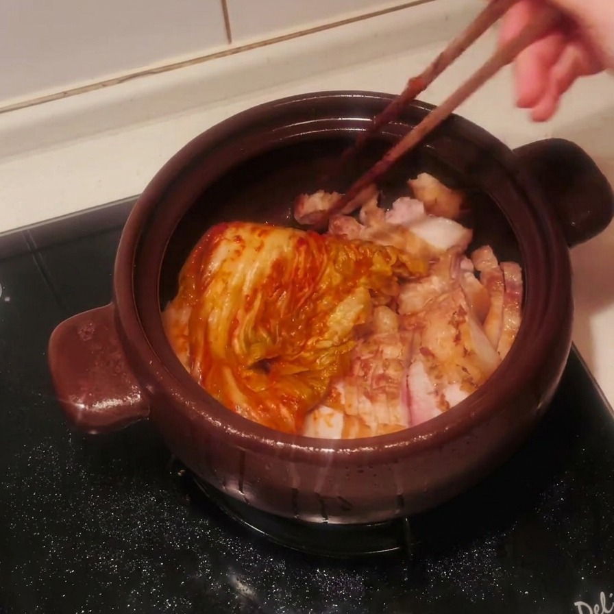 Una pieza entera de kimchi rojo sazonado colocándose en la olla sobre la panceta con palillos de madera.