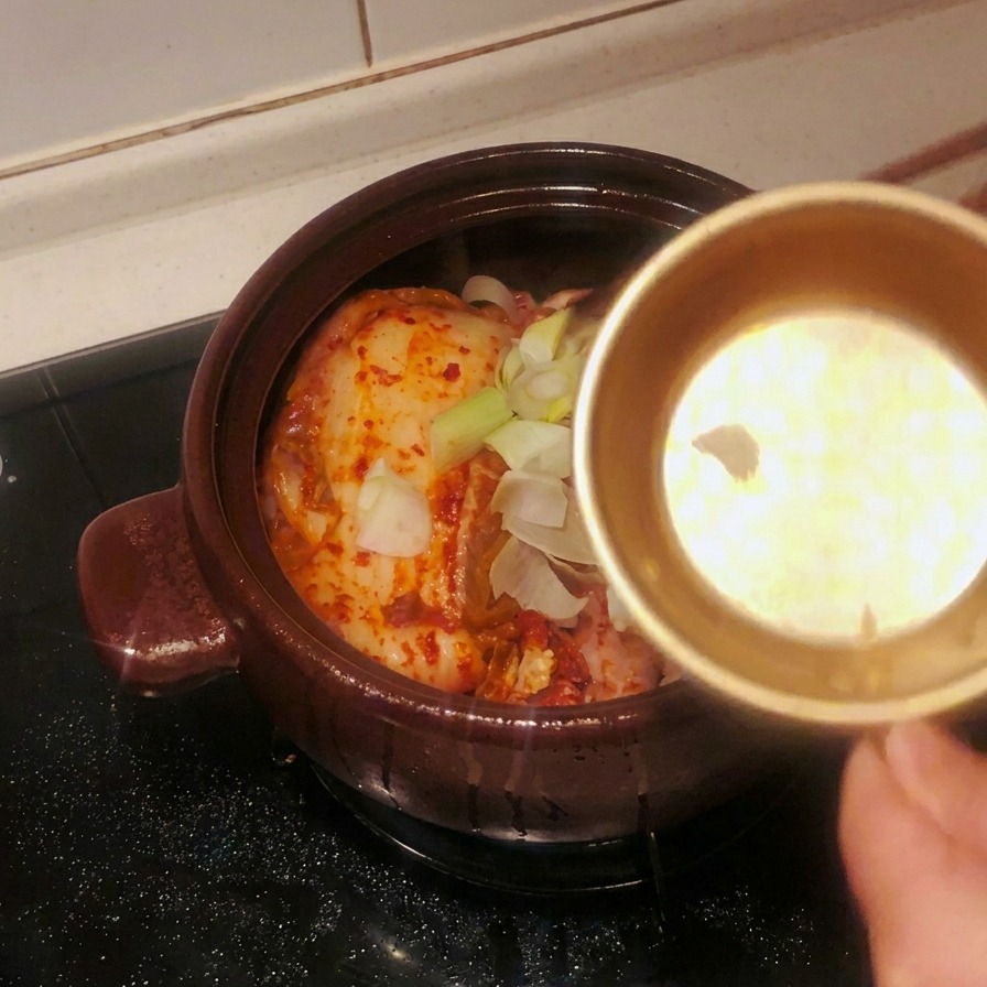 Un bol de metal vertiendo ajo laminado y segmentos grandes de cebolleta verde sobre el kimchi en la olla.