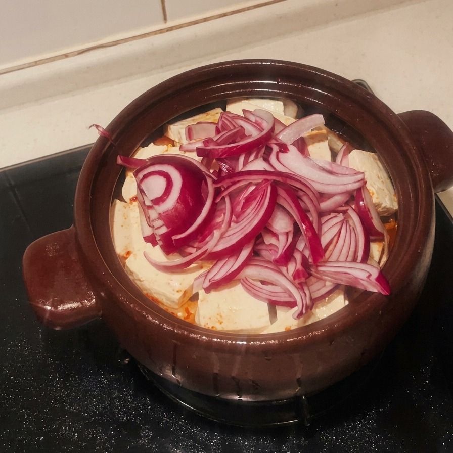 Una pila de cebolla roja laminada cubriendo la capa de tofu blanco en la olla de barro.