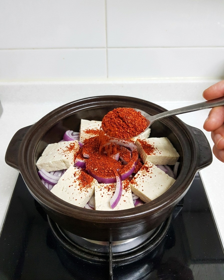 Una cuchara de metal colocando chile rojo fino en polvo sobre el tofu y las cebollas en la olla.