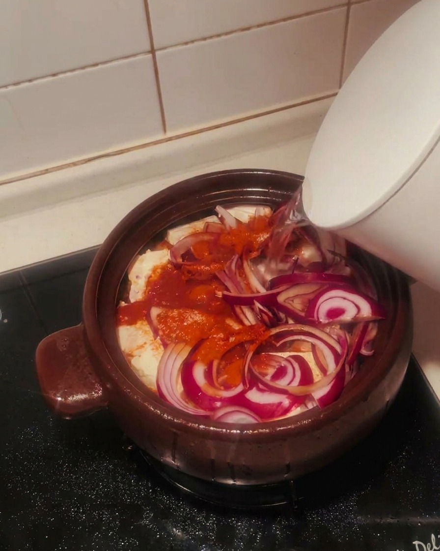 Agua clara vertiéndose desde una tetera blanca en la olla de barro llena de kimchi, tofu, cebolla y especias.