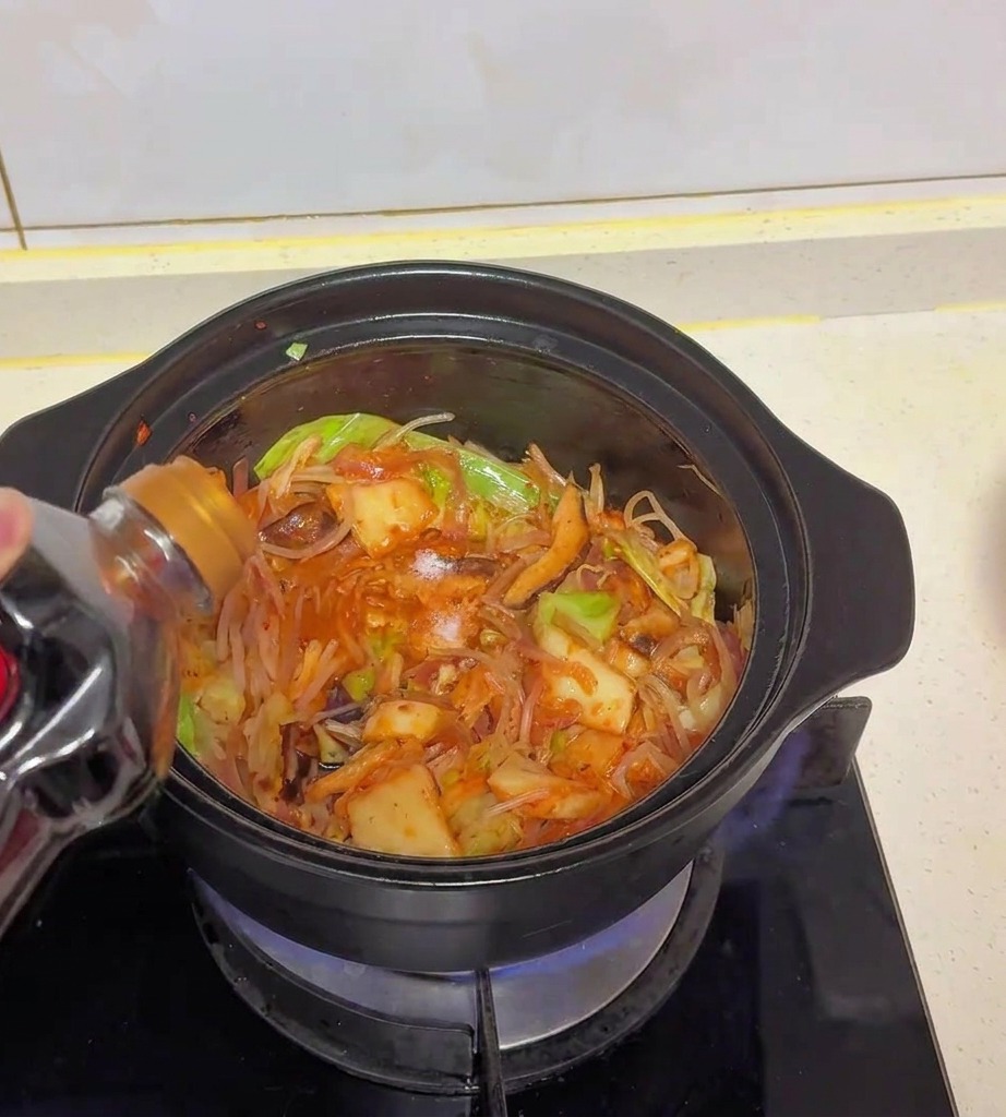 Salsa de soja clara vertiéndose desde una botella de plástico oscura en una olla llena de repollo, kimchi y champiñones.