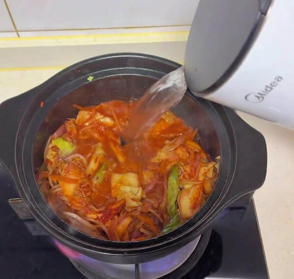 Agua caliente vertiéndose desde un hervidor eléctrico blanco en la olla negra con kimchi y verduras.