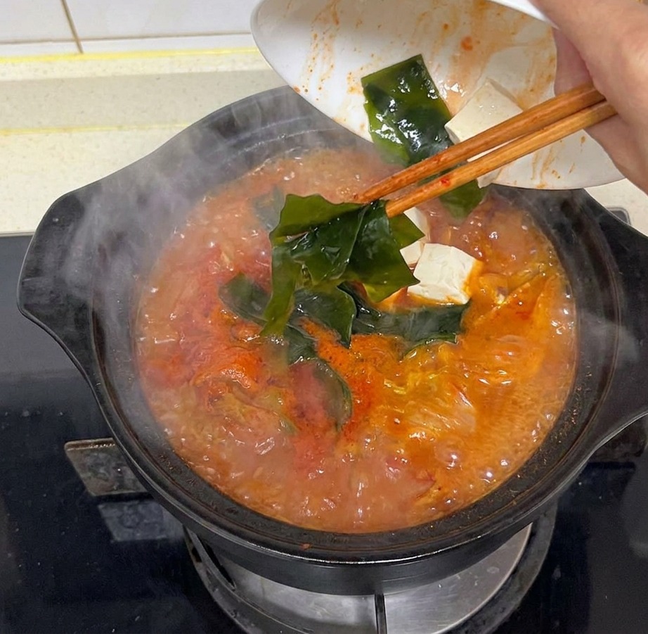 Una mano vertiendo trozos de alga kelp verde oscuro en una olla humeante de sopa de kimchi roja.