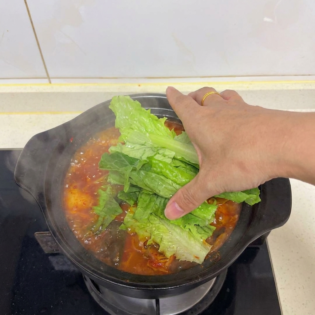 Una mano colocando hojas de lechuga vibrantes sobre una olla de sopa de kimchi y tofu.
