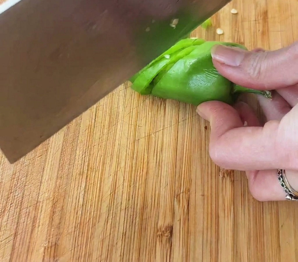 Primer plano de una mano usando un hacha de cocina para picar un chile verde brillante en aros pequeños sobre una tabla de madera.