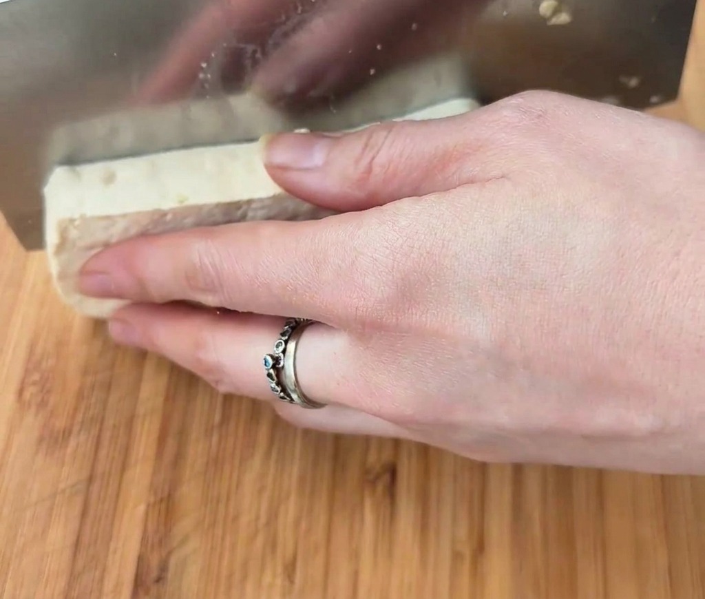 Un bloque rectangular de tofu blanco siendo cortado en cubos con un hacha de metal sobre una superficie de madera.