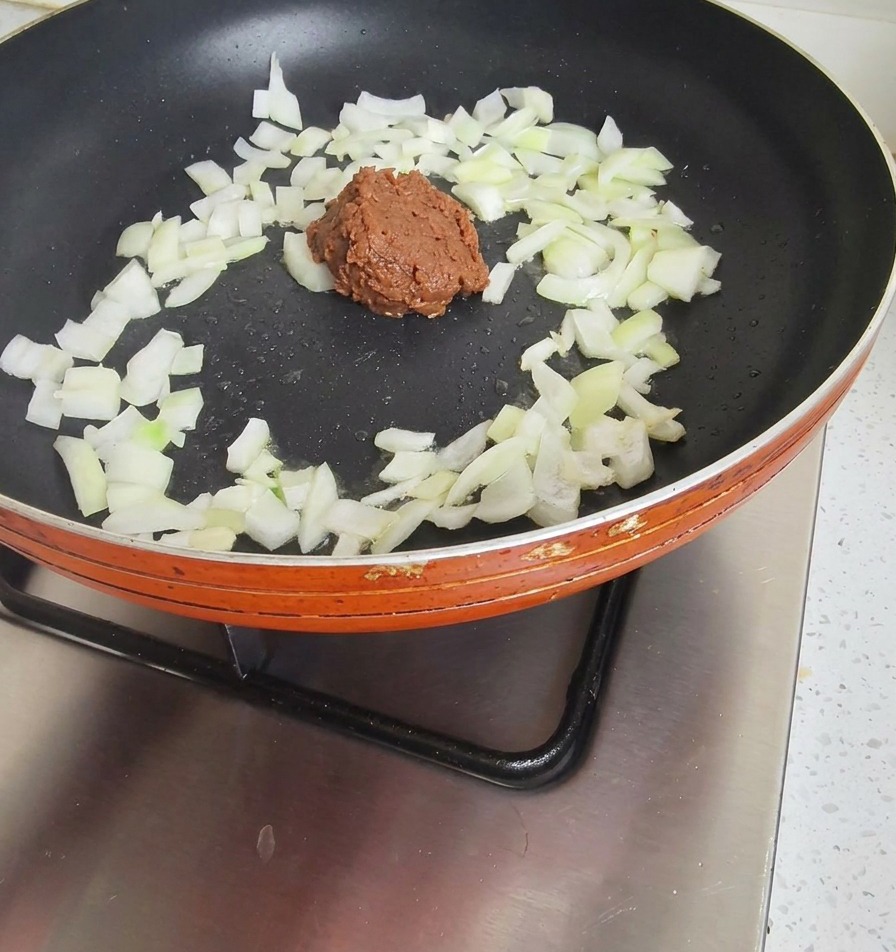 Una porción de pasta de soja espesa y marrón añadida a una sartén con cebollas parcialmente pochadas.