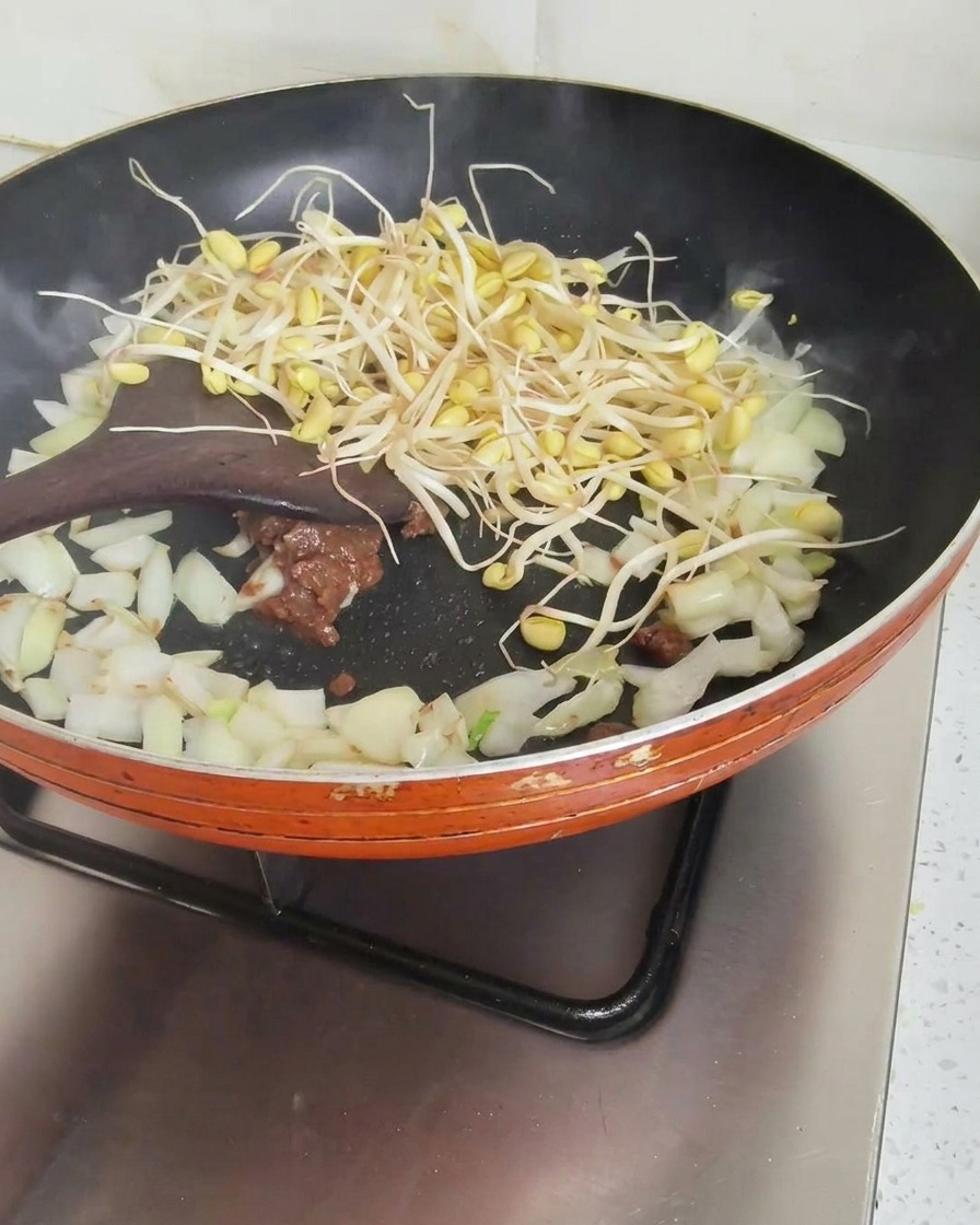 Una espátula de madera removiendo brotes de soja frescos, cebolla picada y pasta de soja en una sartén naranja.