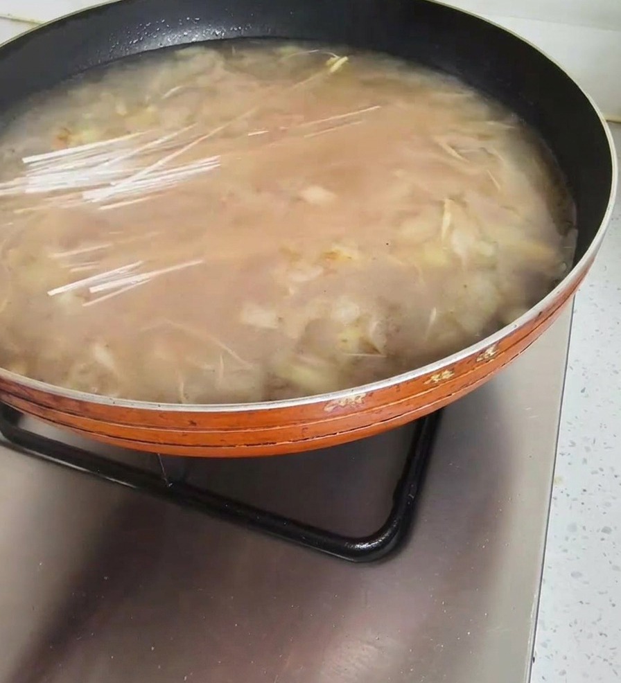 Un puñado de fideos secos y traslúcidos siendo colocados en una sartén con sopa coreana de pasta de soja hirviendo.