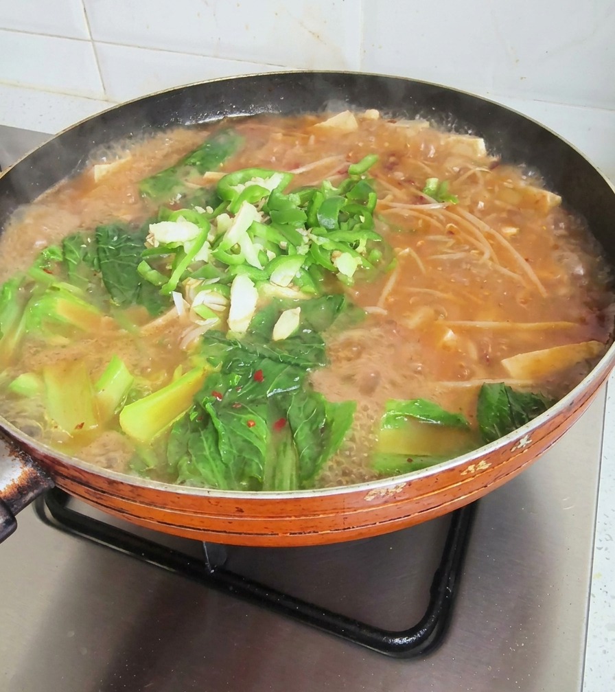 Una sartén burbujeante de sopa coreana cubierta con verduras frescas, chiles picados y ajo picado.