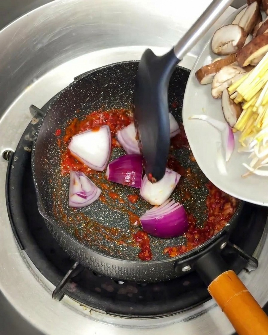 Cebolla morada y setas siendo añadidas a la base de especias rojas en la sartén.