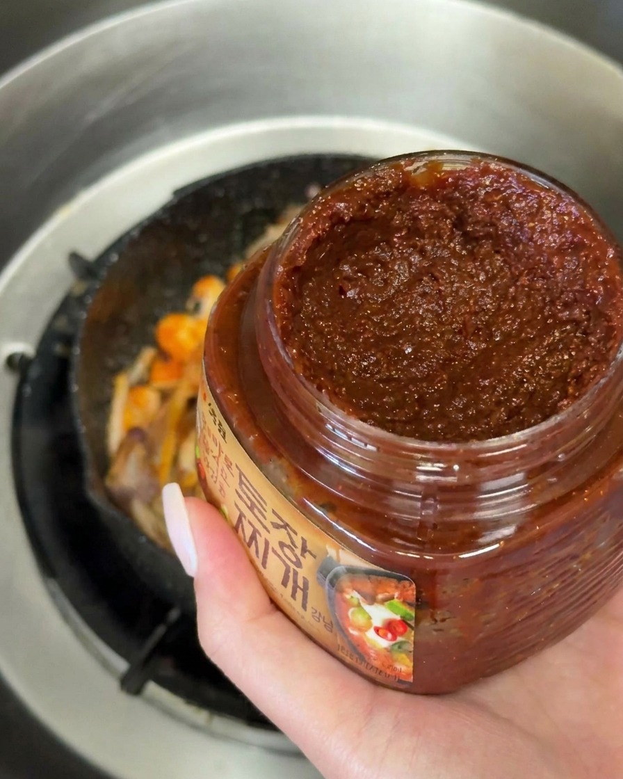 Una mano vertiendo una cucharada de pasta de soja coreana oscura sobre las verduras salteadas.