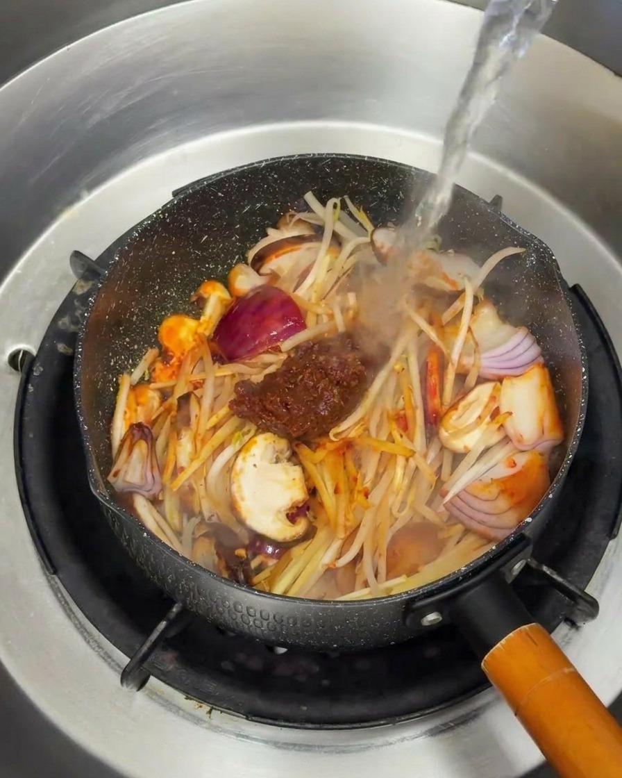 Un chorro de agua hirviendo cayendo sobre las verduras y la pasta de soja en la sartén.