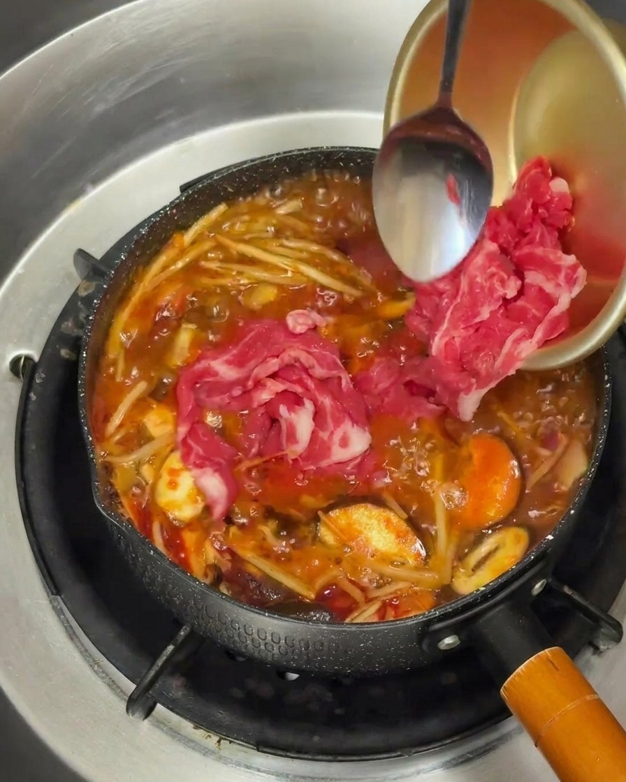 Láminas de ternera cruda siendo añadidas a una olla con caldo rojo burbujeante.
