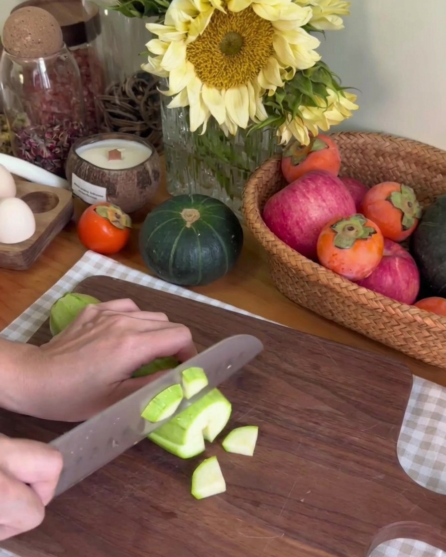 Una persona usando un cuchillo de cocina para cortar un calabacín verde en trozos pequeños sobre una tabla de cortar de madera oscura