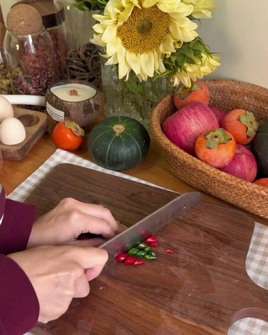 Una persona usando un cuchillo para cortar pequeños chiles verdes y rojos en anillos finos sobre una tabla de madera.