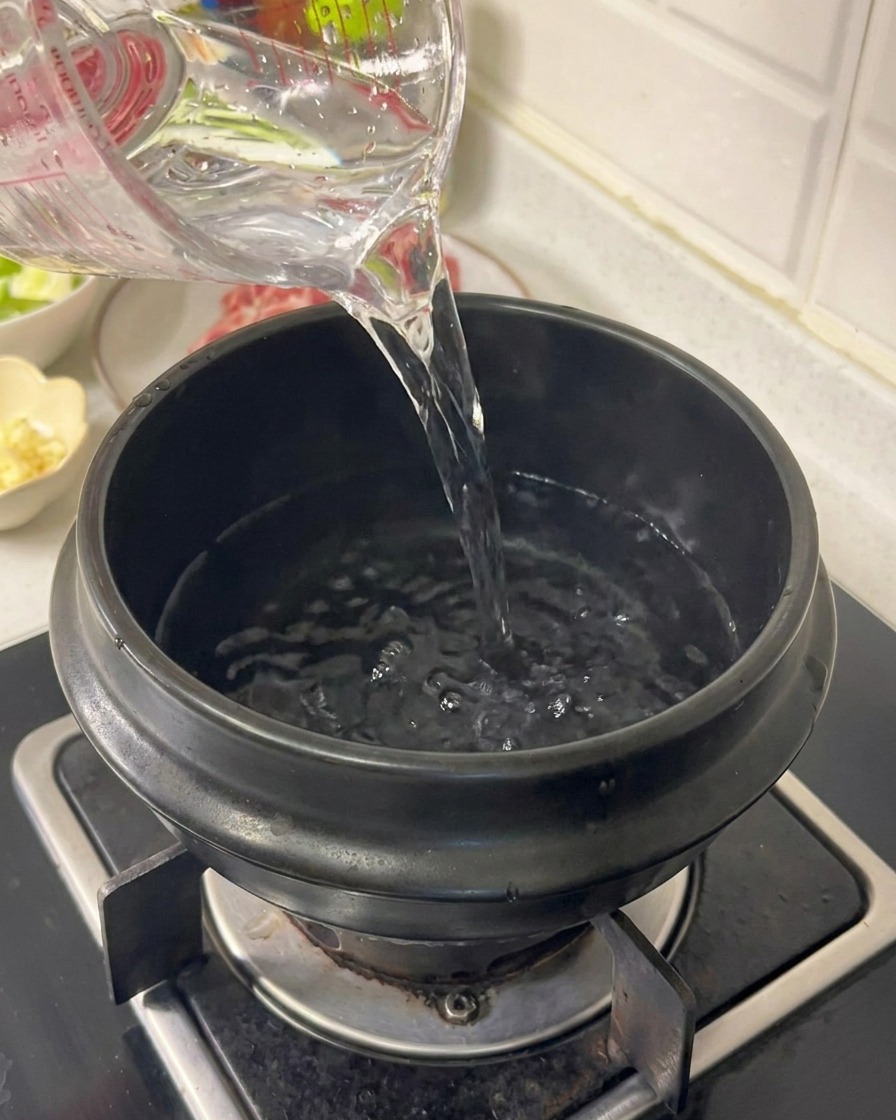 Agua vertiéndose desde una jarra medidora transparente en una olla de cerámica negra sobre una estufa de gas.