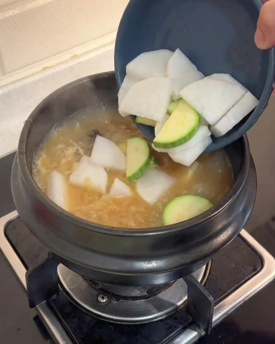 Rodajas de nabo blanco y calabacín verde siendo vertidas desde un bol azul en una olla hirviendo de caldo marrón.