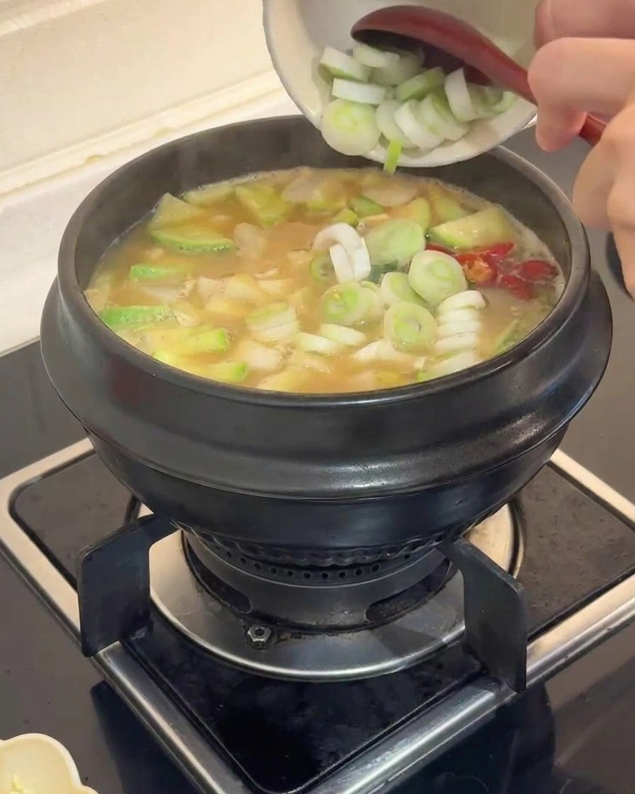 Puerros verdes troceados siendo añadidos desde un pequeño bol blanco a una olla de piedra burbujeante con estofado de verduras.
