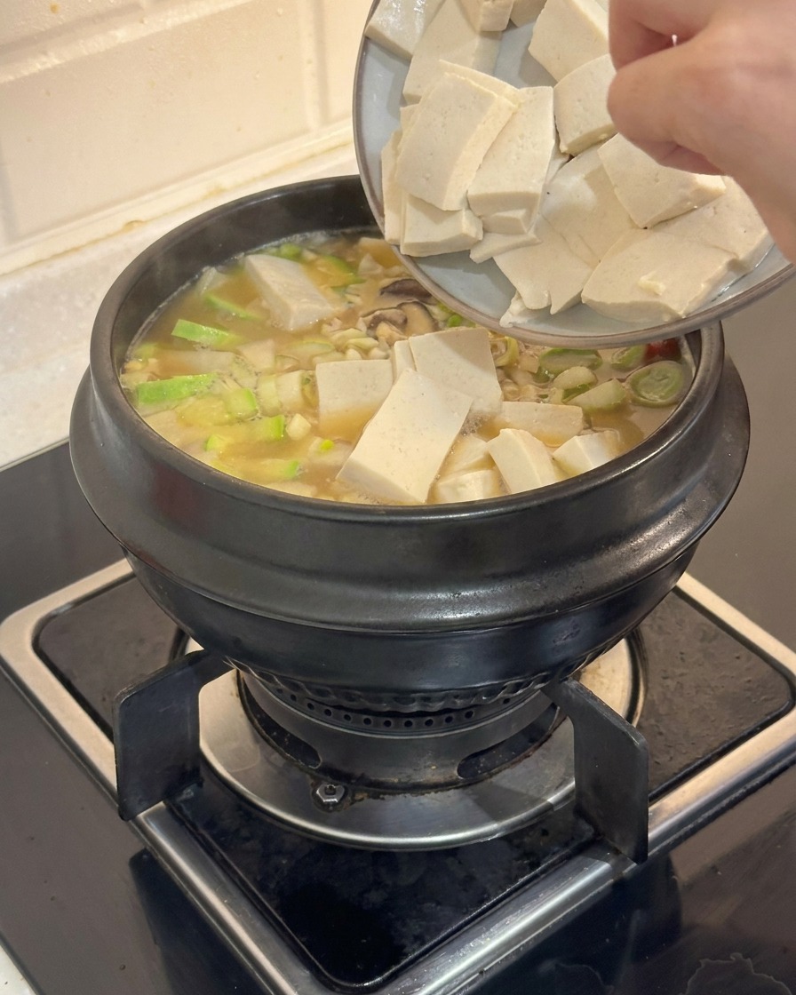 Una mano deslizando cubos rectangulares de tofu blanco desde un plato hacia una olla humeante de estofado coreano.