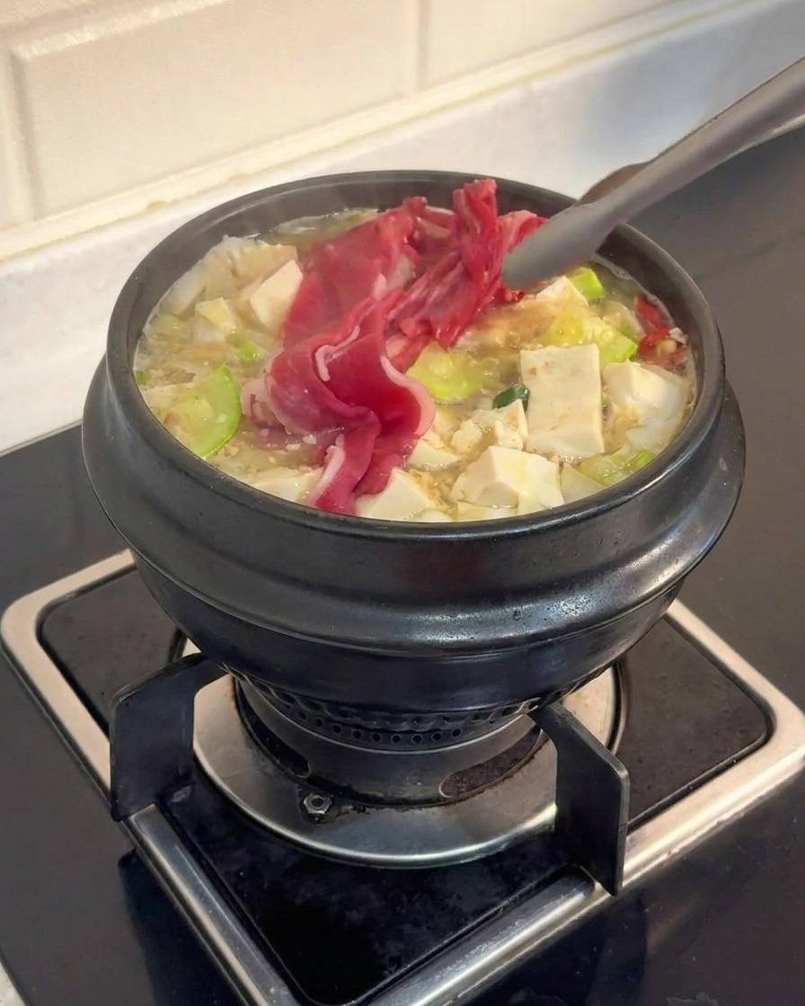 Pinzas metálicas colocando finas láminas de ternera cruda en una olla llena de tofu y verduras en un caldo burbujeante.