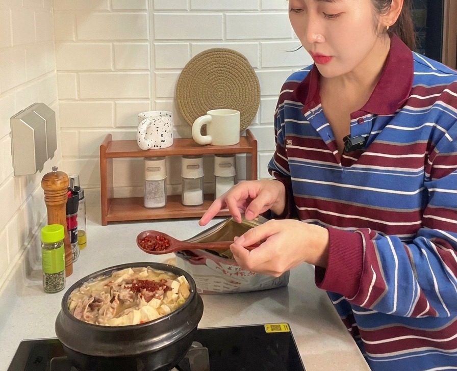 Una cuchara de madera añadiendo chile rojo en polvo brillante a una olla burbujeante de estofado coreano con ternera, tofu y calabacín.