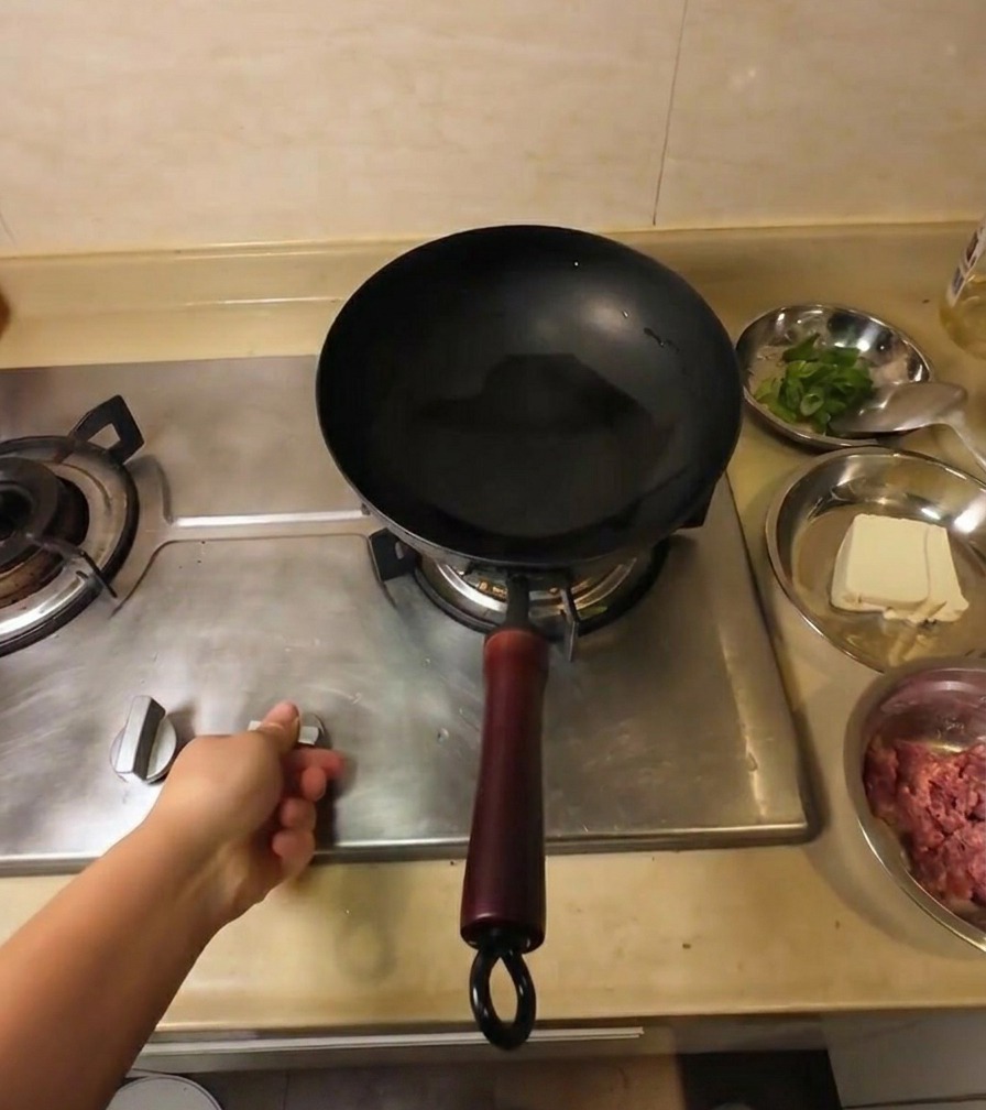 Una mano ajustando el mando de la estufa de gas bajo un wok negro con aceite de cocina transparente.