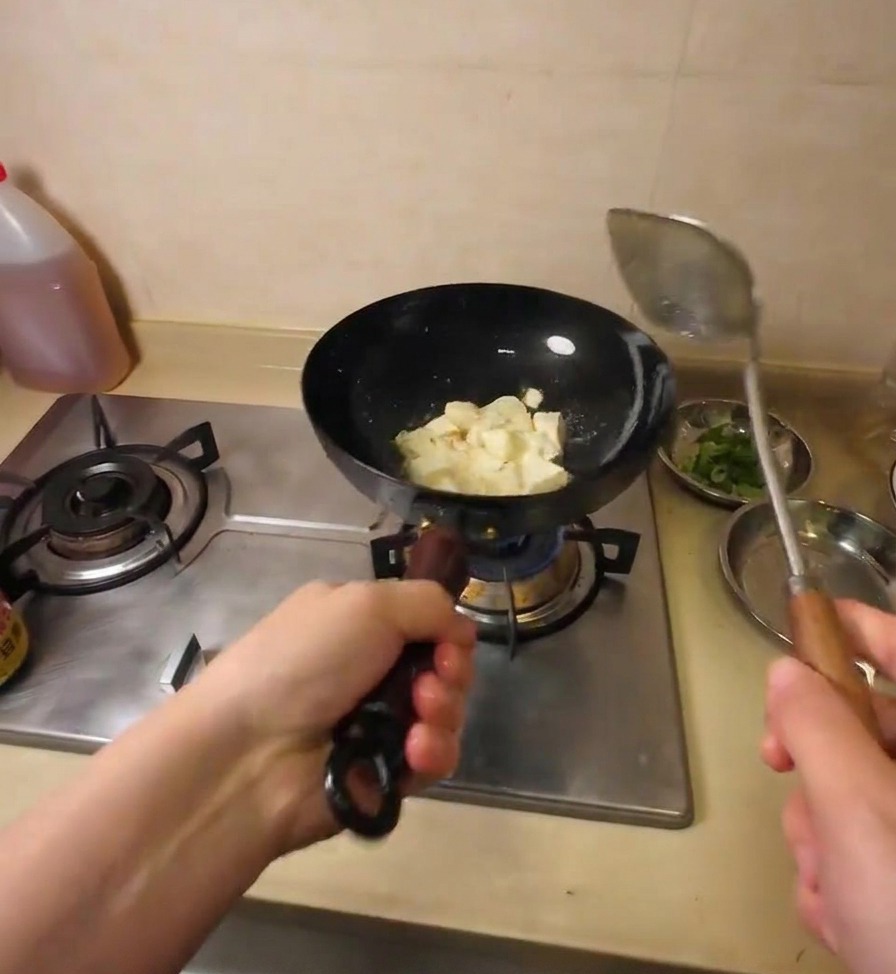 Una espátula metálica rompiendo un bloque de tofu en trozos más pequeños mientras se dora en aceite caliente.