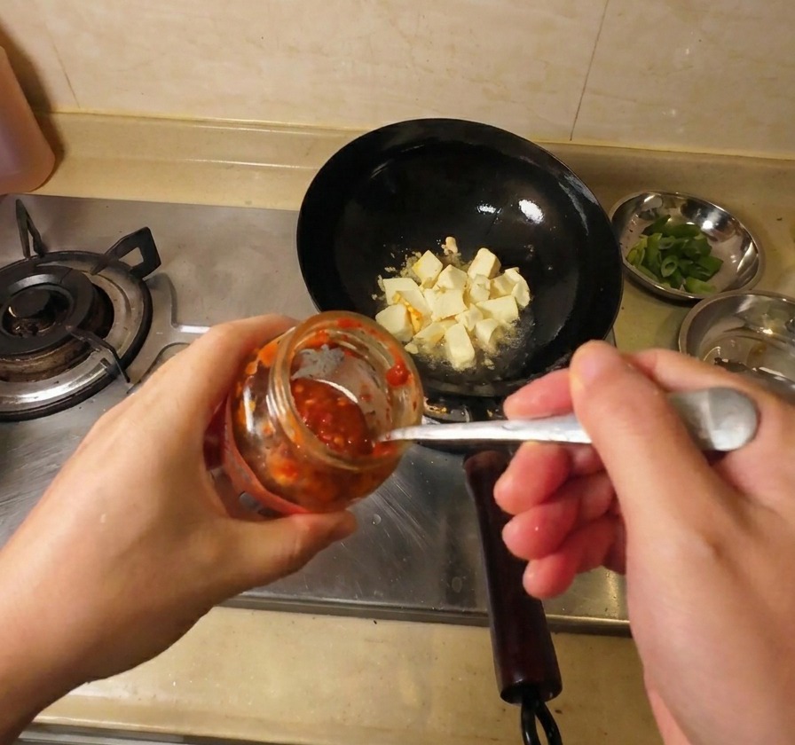 Primer plano de una mano usando una cuchara metálica para añadir salsa de chile roja desde un frasco de vidrio a un wok con tofu dorado.