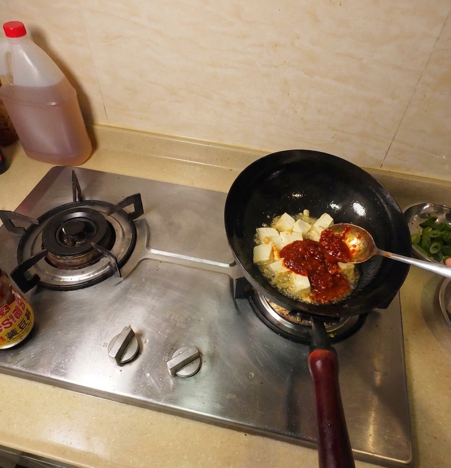 Pasta de soja de color marrón oscuro en una cuchara metálica añadiéndose al wok junto al tofu frito y la salsa de chile.