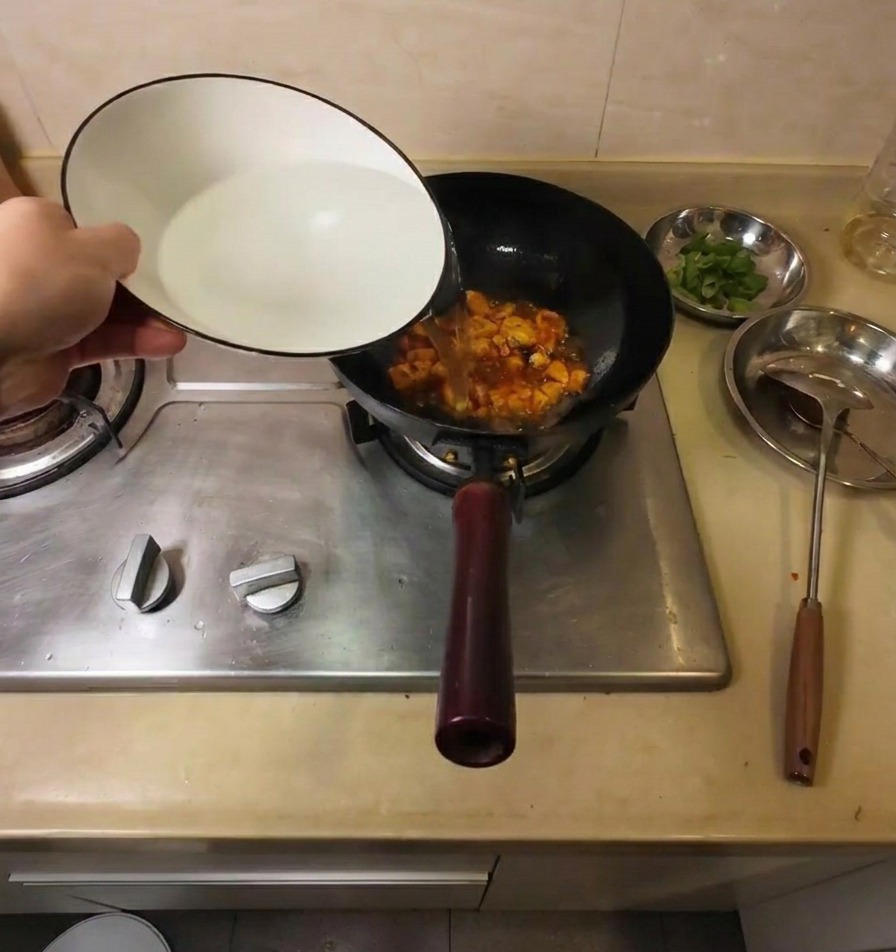 Agua caliente vertiéndose desde un cuenco de cerámica blanca en un wok negro con tofu y condimentos rojos.