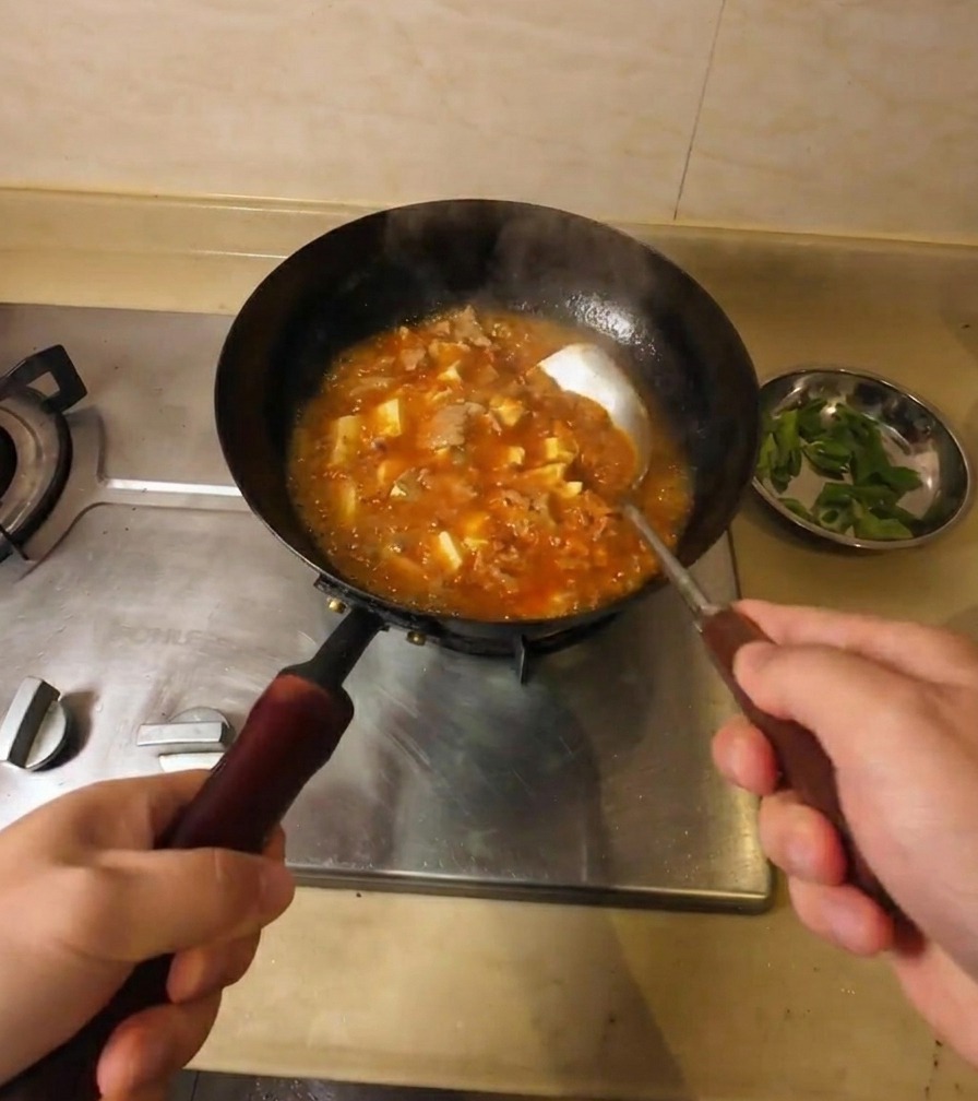 Una persona removiendo una sopa espesa de color naranja rojizo con trozos de tofu y ternera en un wok sobre la estufa.