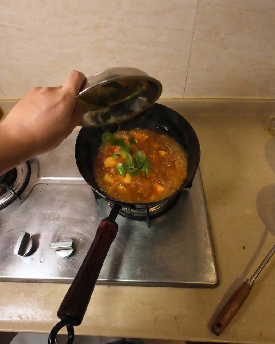 Una mano vertiendo cebolletas frescas picadas desde un pequeño cuenco de metal a un wok con sopa de ternera y tofu hirviendo.