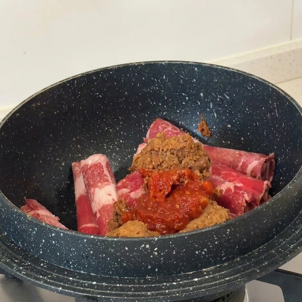 Pasta de soja marrón y pasta de chile roja sobre láminas de ternera cruda en una olla oscura.